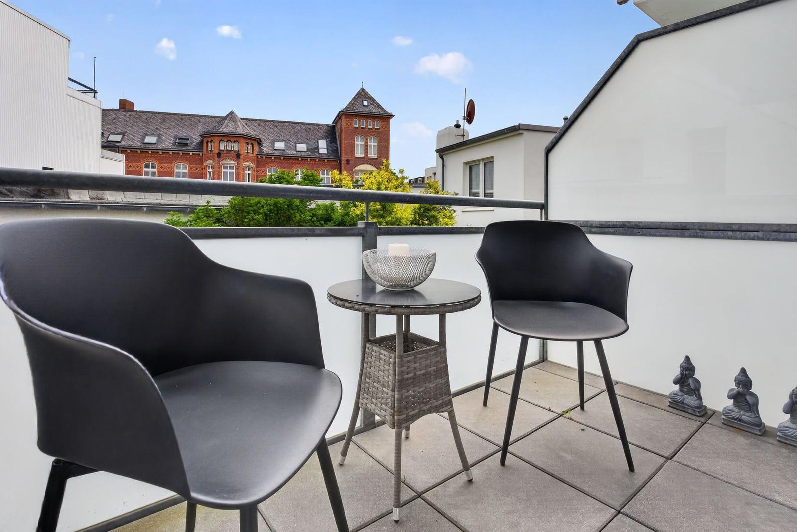Terrasse mit zwei Stühlen und Tisch. Im Hintergrund ein rotes Gebäude.