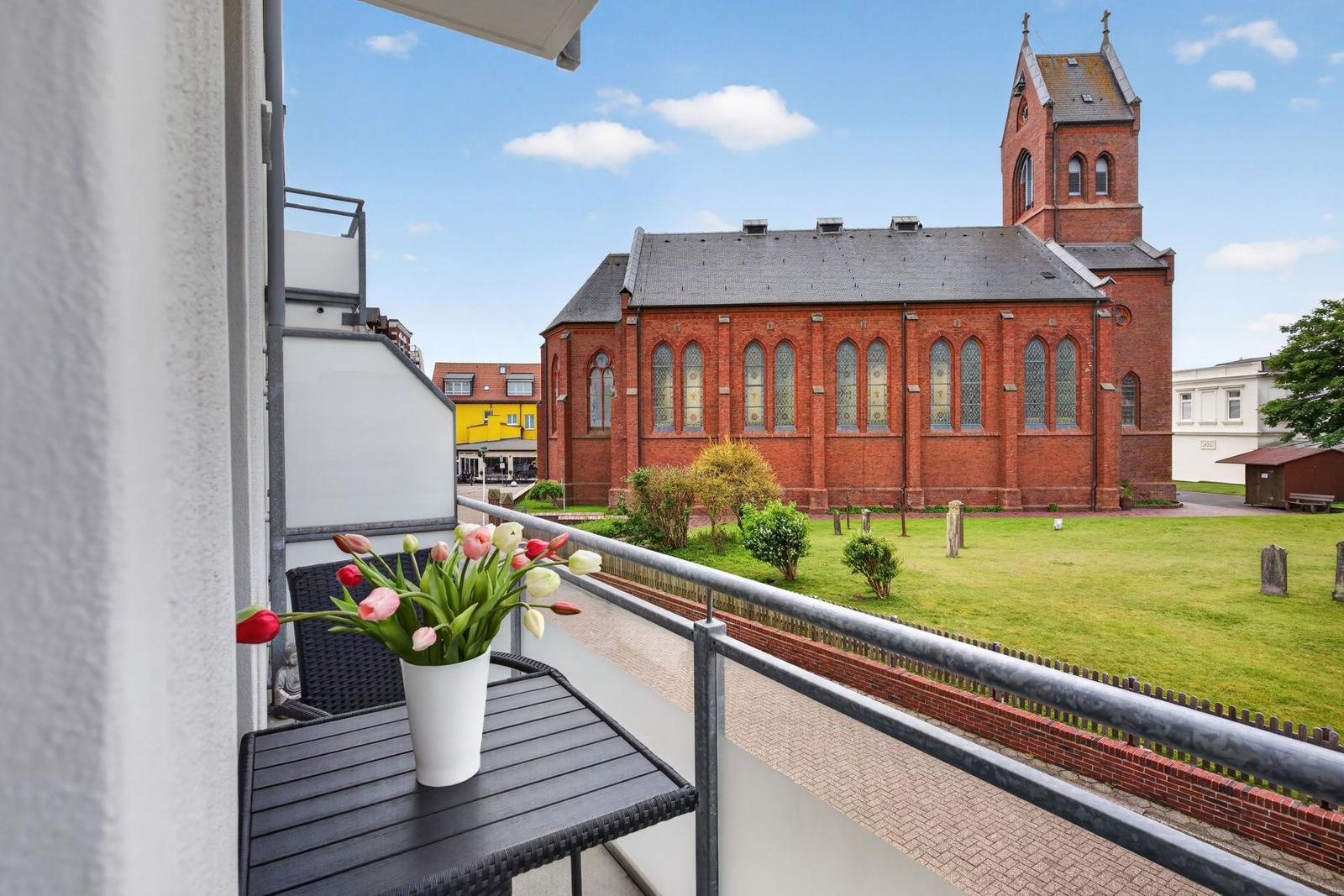 Balkon mit Blumen und Blick auf eine rote Backstein-Kirche mit Grünfläche.