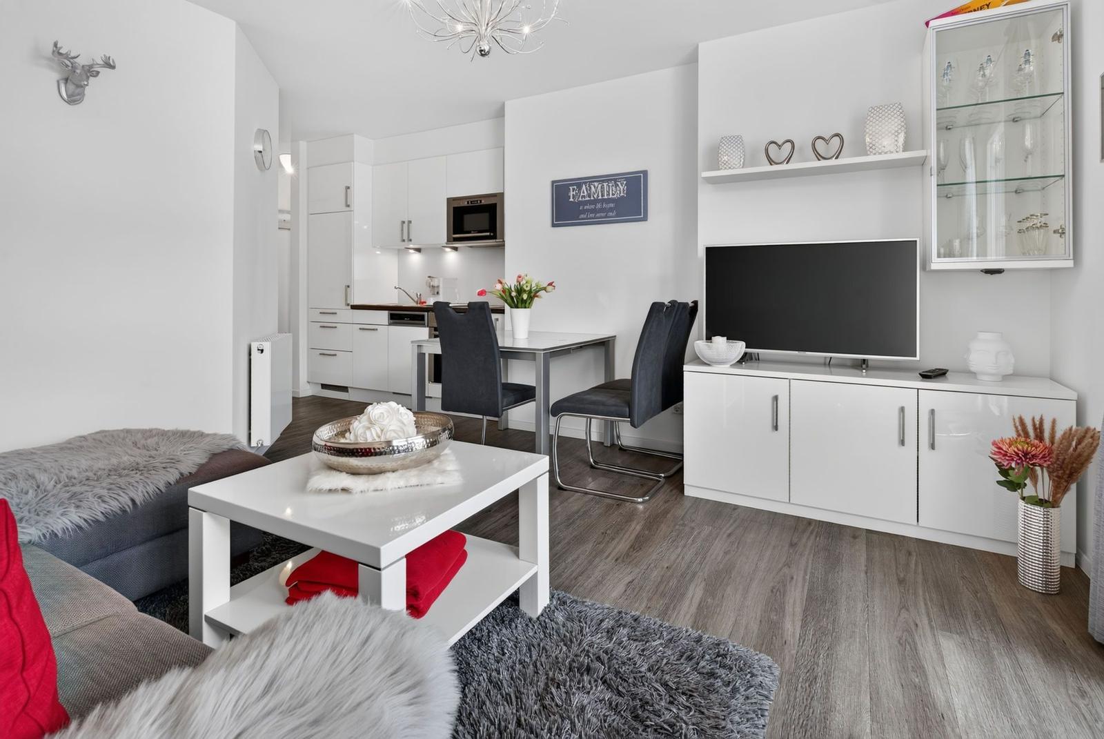 Wohnzimmer mit TV, Esstisch und Küche im Hintergrund. Weiße Möbel und Holzboden.