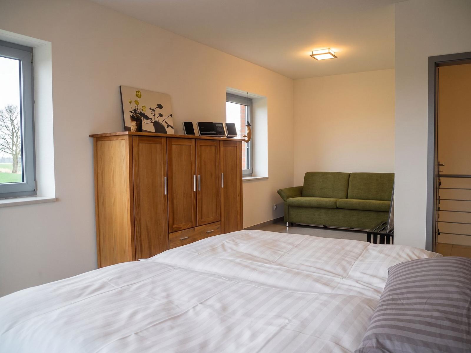 Schlafzimmer mit Holzschrank, Fenster und Bett. Auf dem Schrank steht ein Bild und ein Radio.