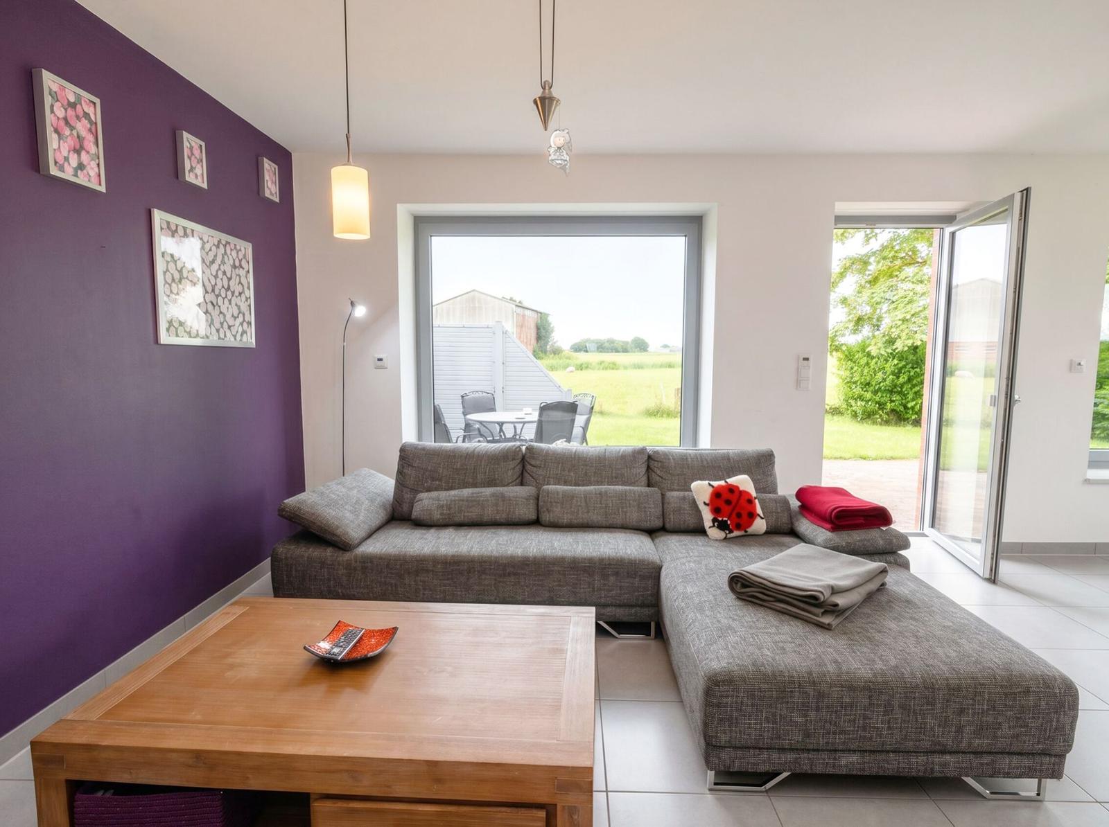 Wohnzimmer mit grauem Sofa, Holztisch und lila Wand. Durchgänge zu Terrasse und Garten.