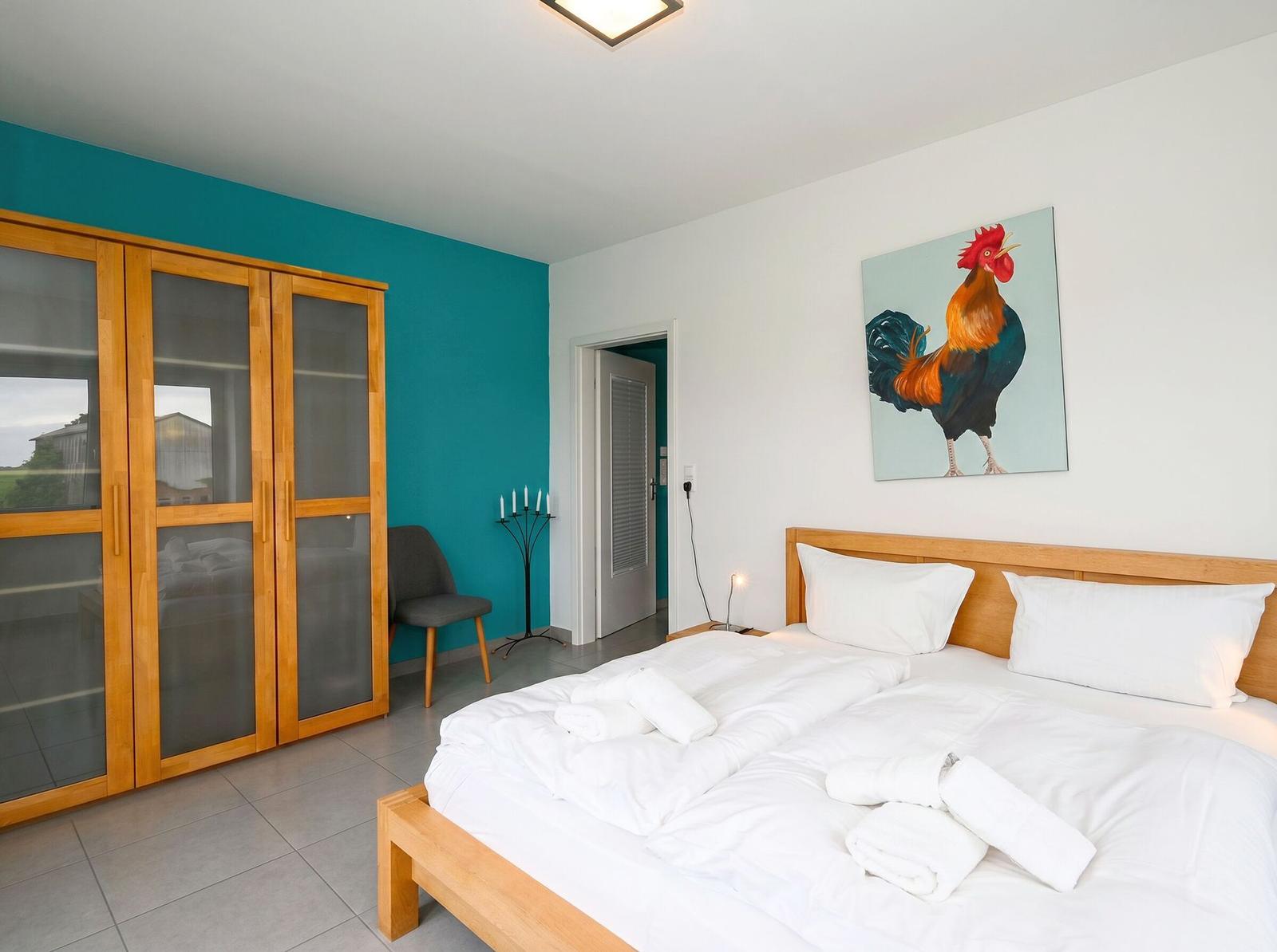 Schlafzimmer mit Bett, Kleiderschrank und Gemälde eines Hahns an der Wand.