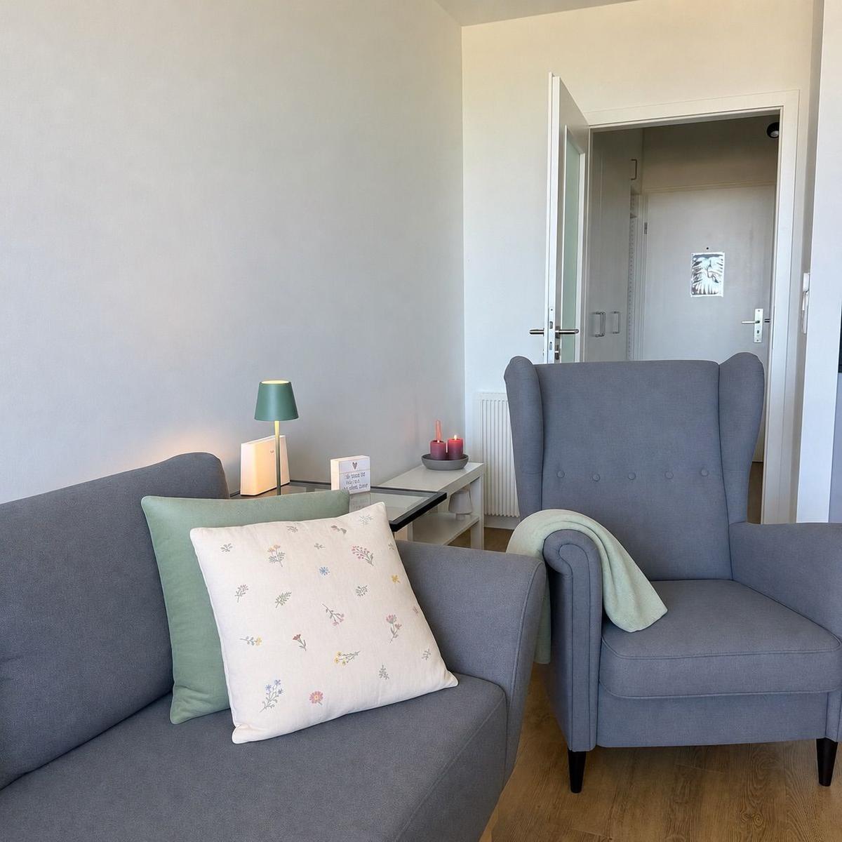 Ein Wohnbereich mit grauem Sofa, Sessel und Beistelltisch. Auf dem Sofa liegen Kissen.