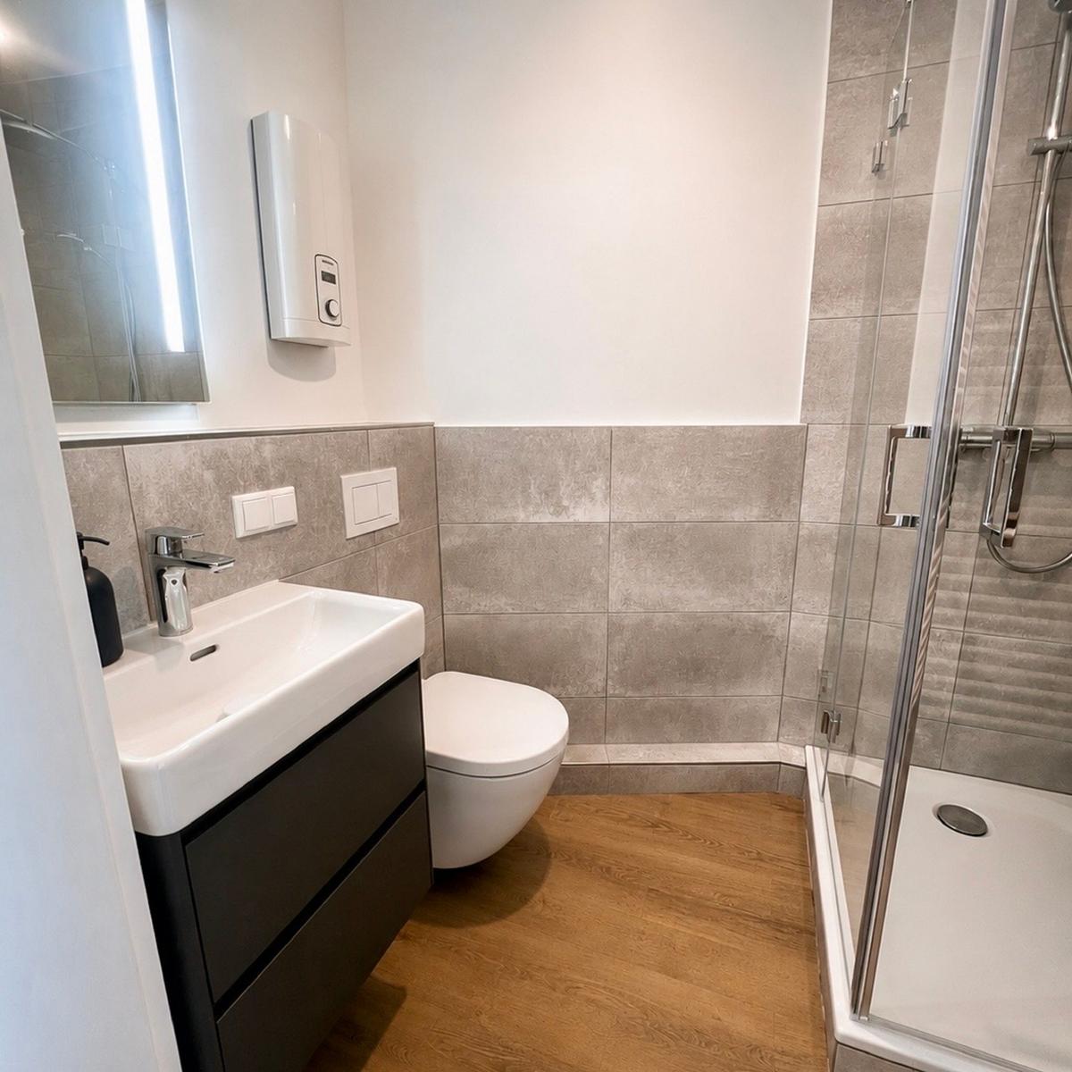 Badezimmer mit Dusche, Waschbecken und Toilette. Holzboden und graue Fliesen.