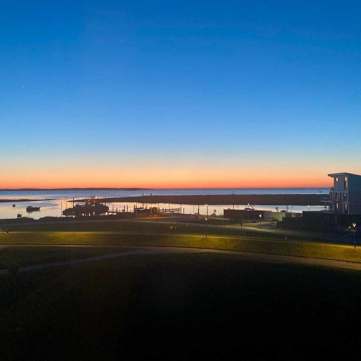 Sonnenuntergang über Wasser mit Booten und modernen Gebäuden im Hintergrund.