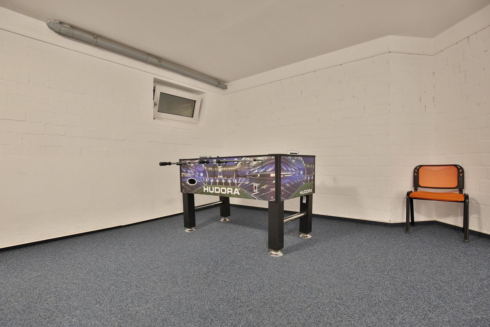 Fussballtisch mit Stadionmotiv, orange Sitzgelegenheit, weiße Wände und grauer Boden.