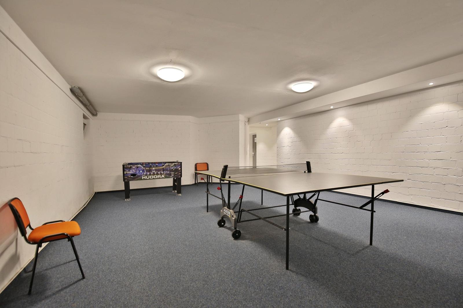 Raum mit Tischtennisplatte und orangefarbenen Stühlen auf blauem Boden.