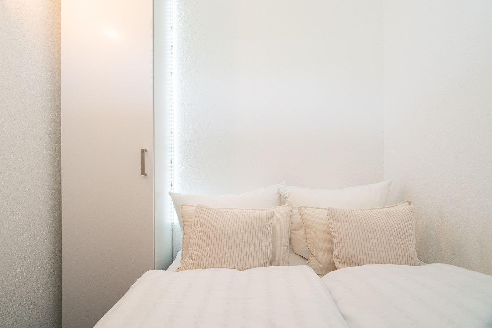 Doppelbett mit Kissen und Bettwäsche in einem hellen Raum mit Fenster und Schrank.