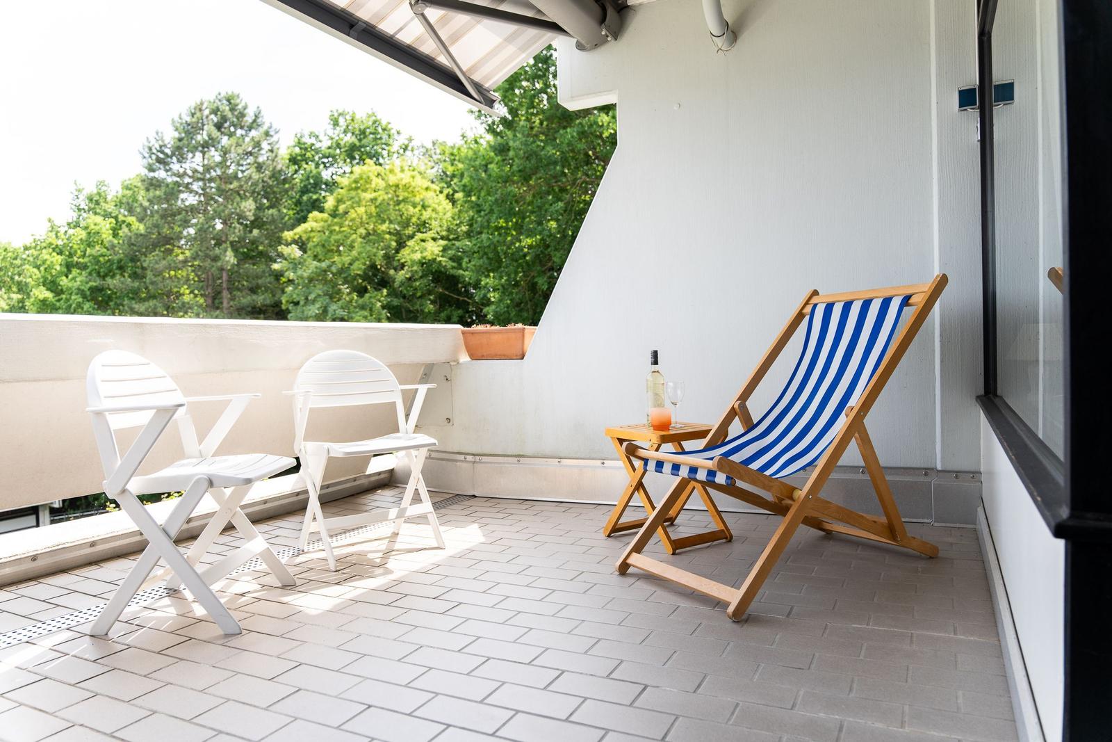 Terrasse mit zwei weißen Stühlen und einem blau-weißen Liegestuhl. Hintergrund: grüne Bäume.