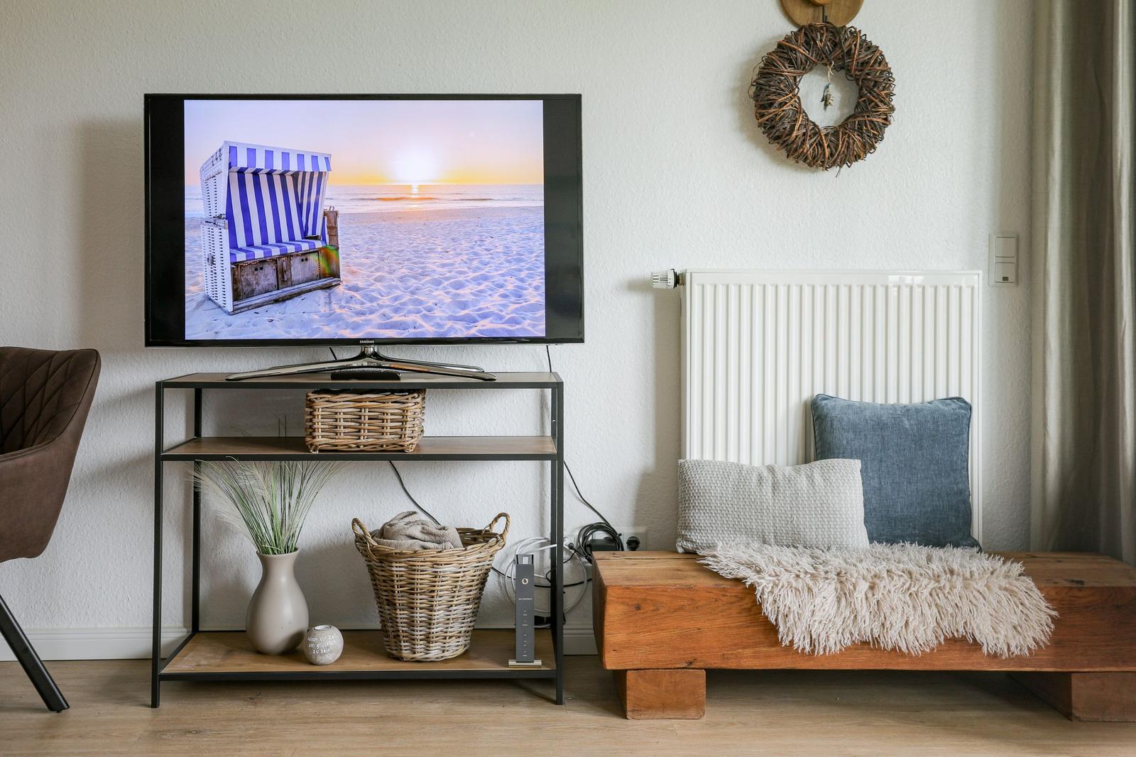 TV auf Ständer mit Körben und Kissen, daneben Holzbank mit Decke.