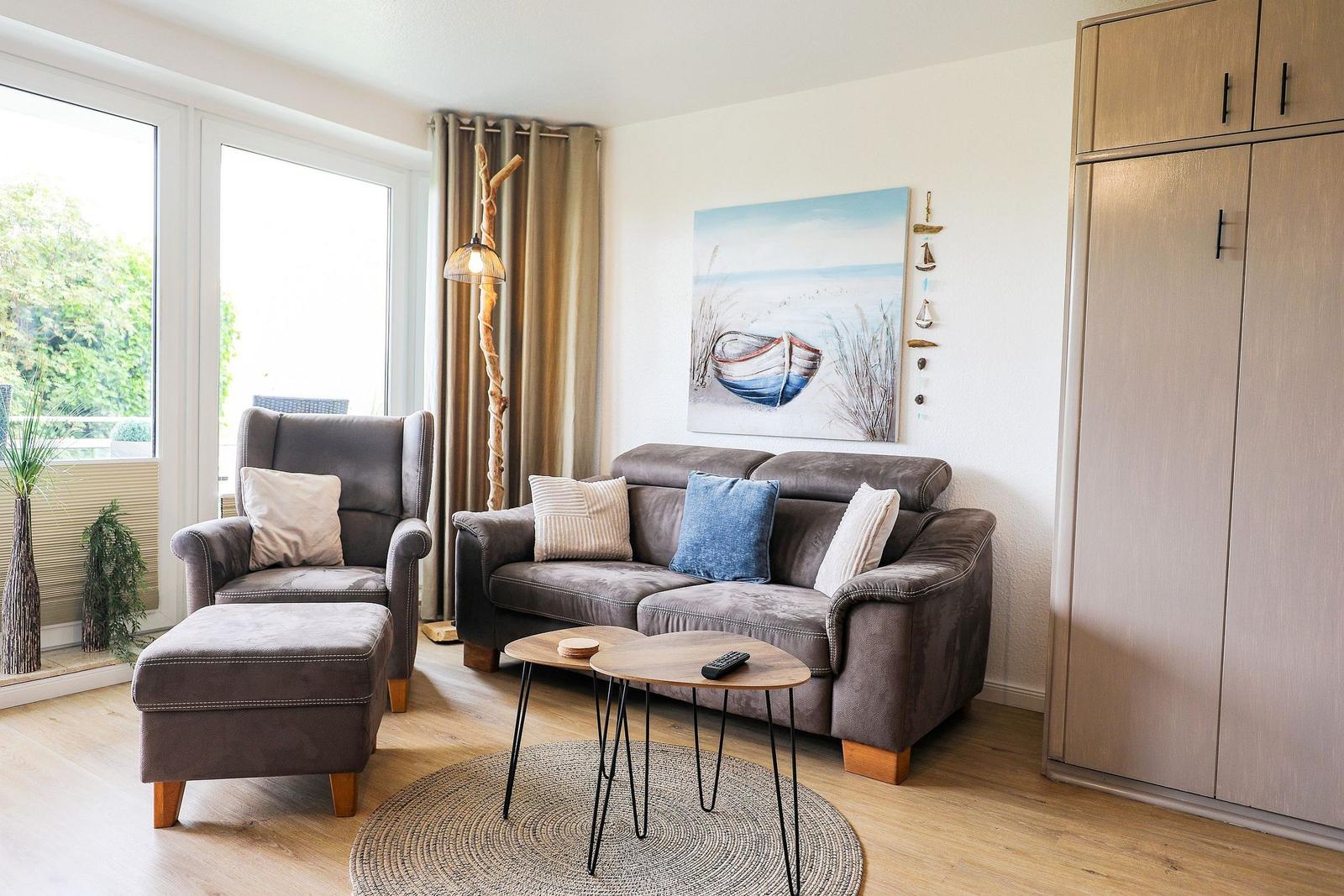 Wohnzimmer mit Sofa, Sessel, Tisch und Wandbild. Holzfußboden und große Fenster.
