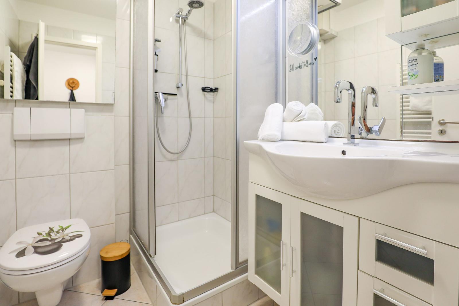 Badezimmer mit Dusche, Waschbecken und Toilette. Weiße Fliesen und moderne Einrichtung.