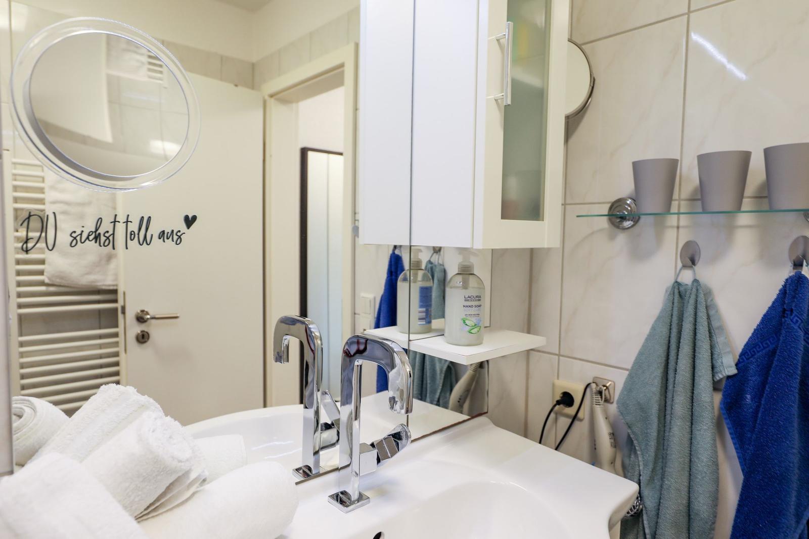 Badezimmer mit Waschbecken, Spiegel, Handtuchhaltern und Duschbereich.