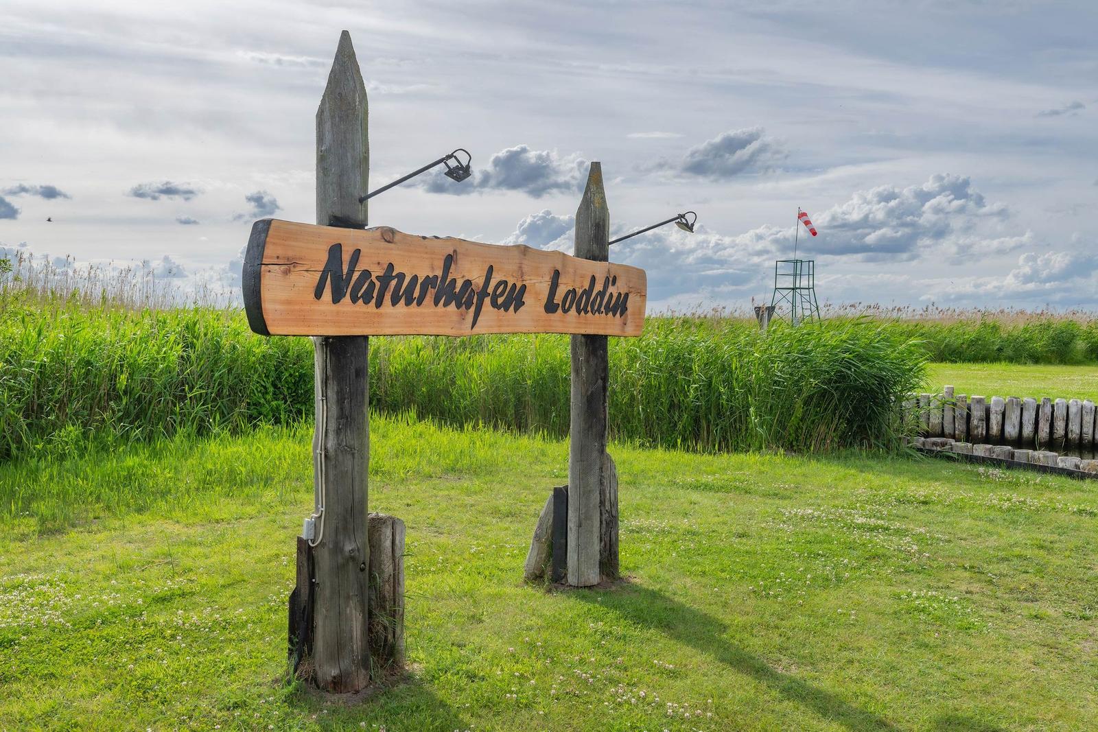 Holzschild mit der Aufschrift Naturhafen Loddin inmitten von Gras und Himmel.