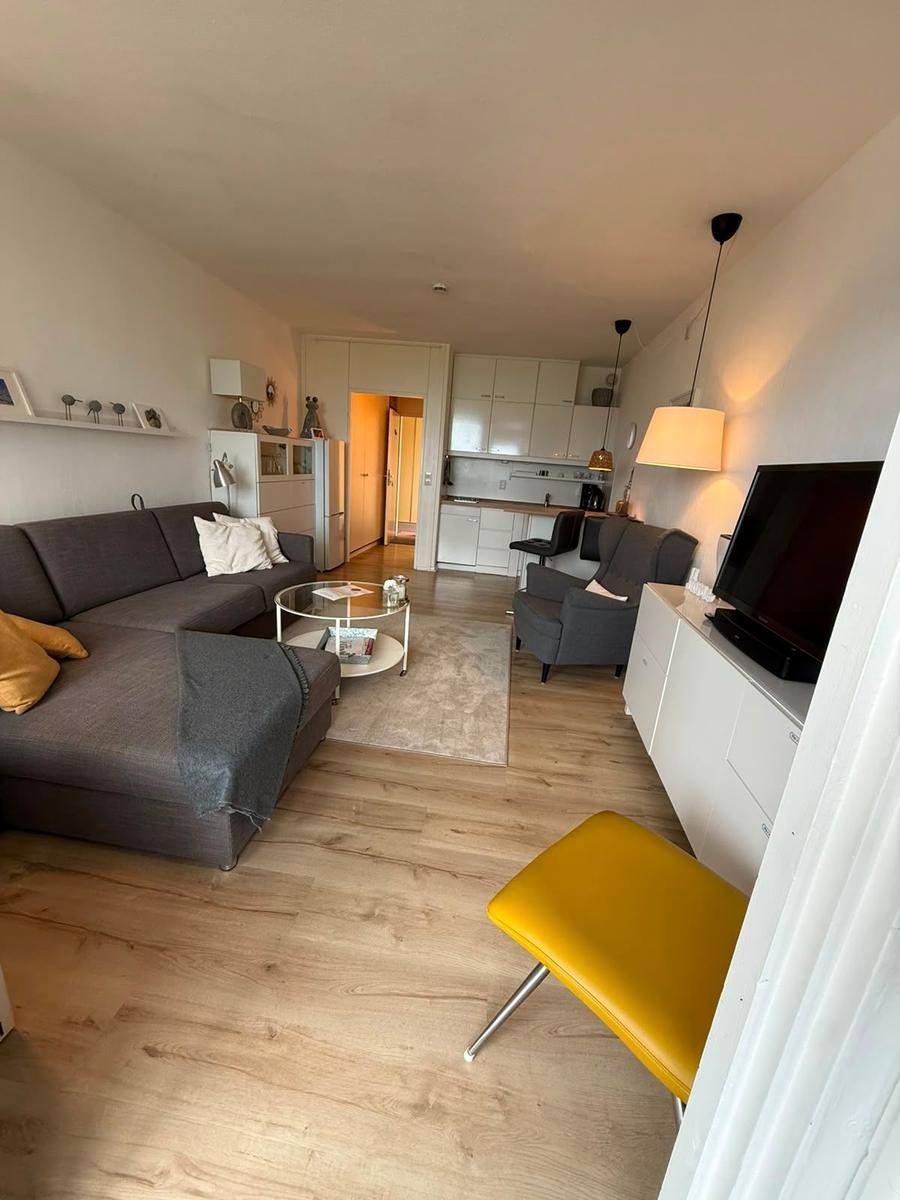 Wohnzimmer mit Sofa, Sessel und Küchenbereich. Holzboden und gelber Stuhl.