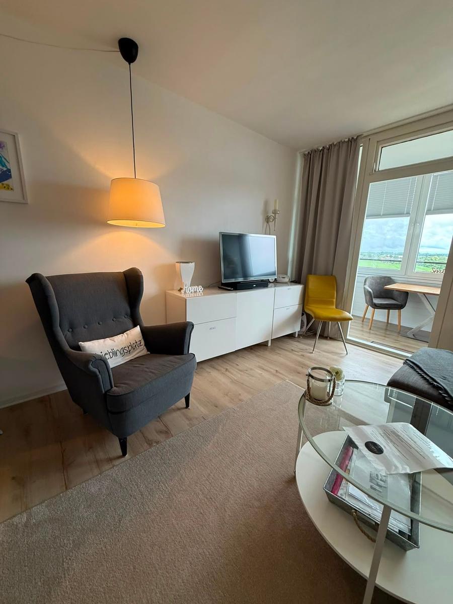 Wohnzimmer mit Sessel, Fernseher und Glas-Tisch. Durchgang zur Terrasse mit Sitzgelegenheit.