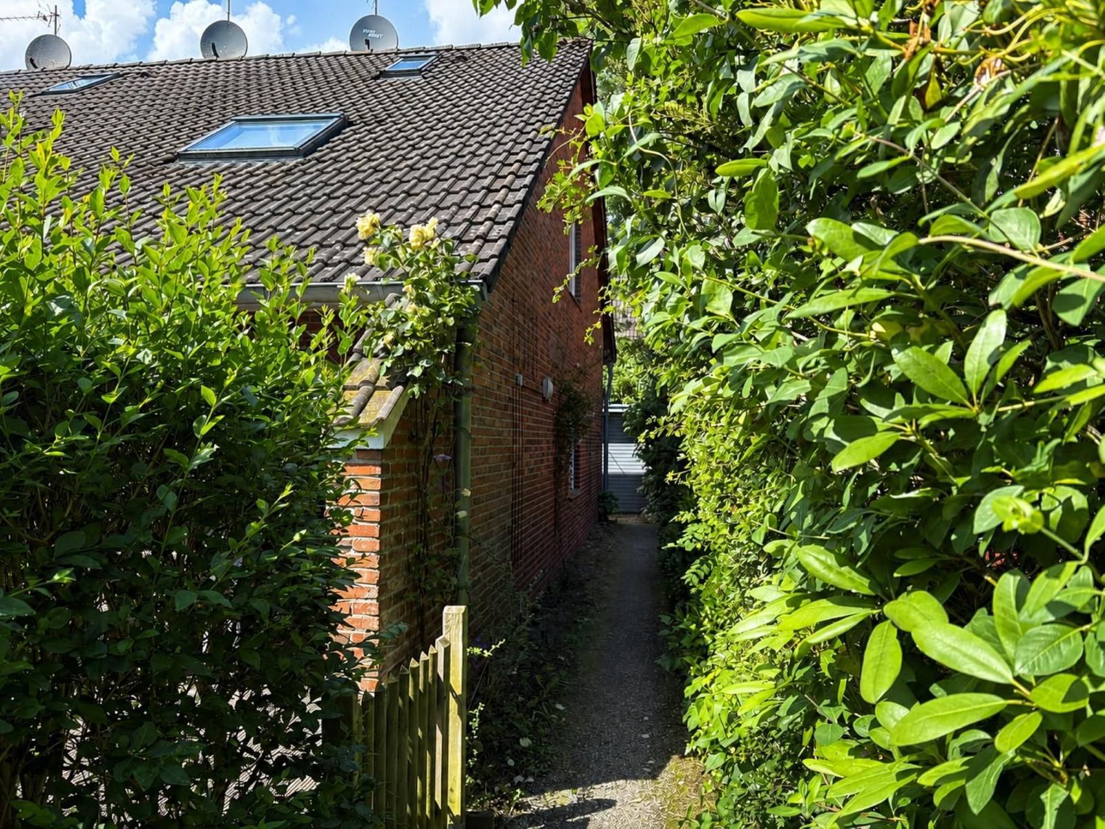 Ein schmaler Pfad führt zwischen grünen Hecken zu einem Backsteinhaus mit Dachfenstern.