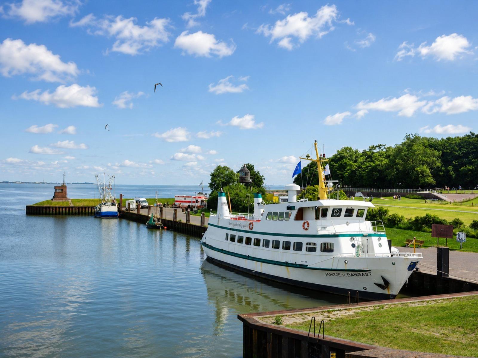 Ein weißes Schiff mit grüner Linie liegt am Hafen. Im Hintergrund sind Bäume und ein kleiner Leuchtturm zu sehen.