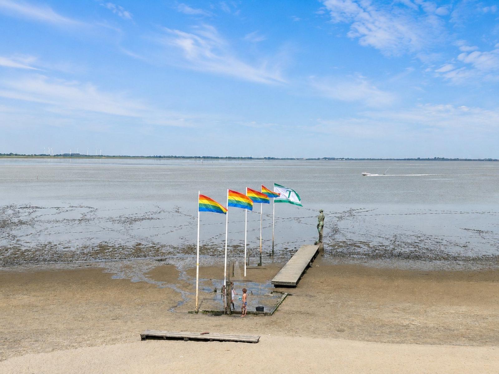 Strand mit Flaggen, Steg und Statue am Wasser.