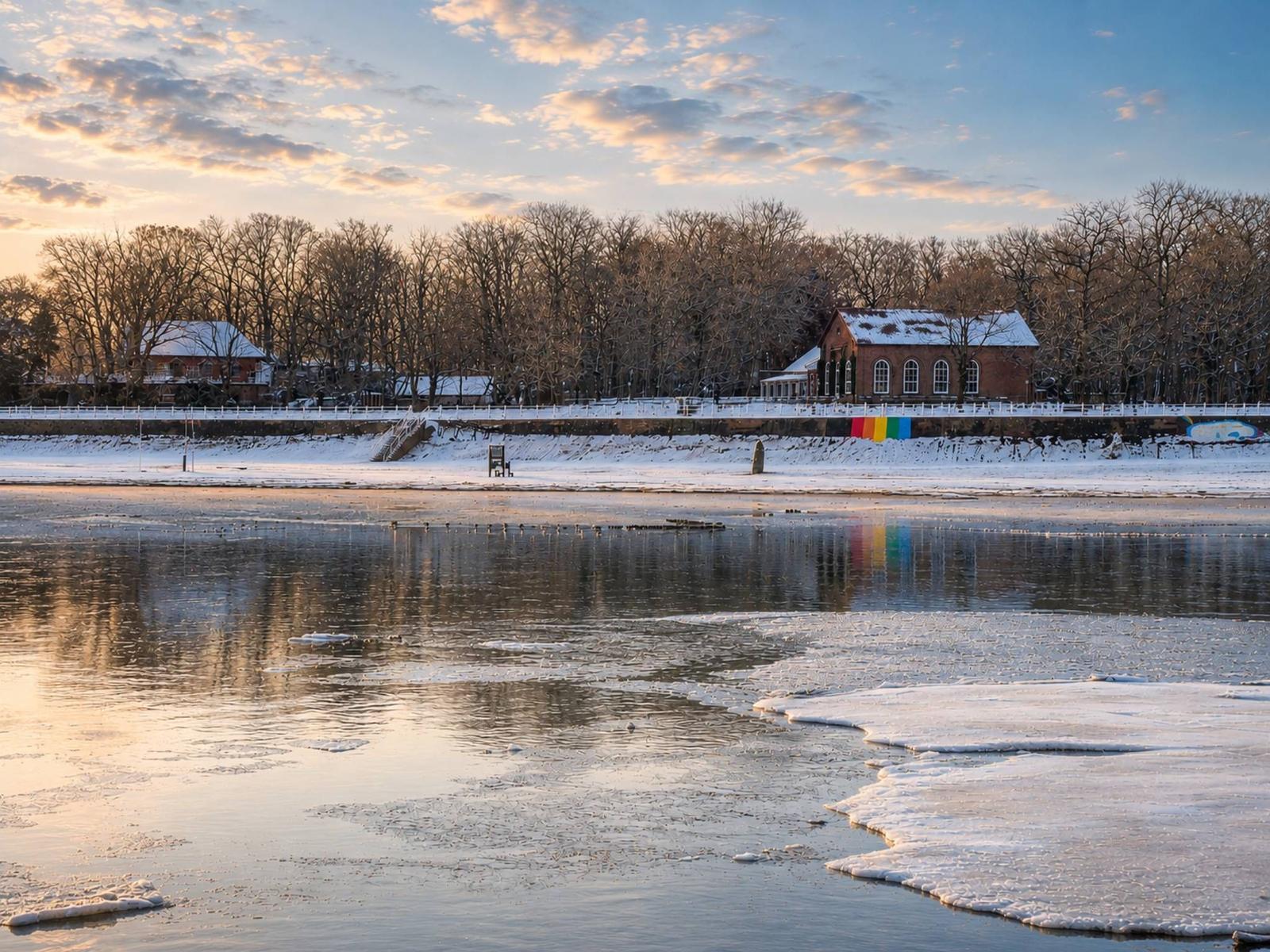 Schneebedeckter Uferbereich mit gefrorenem Wasser und rotem Gebäude im Hintergrund.