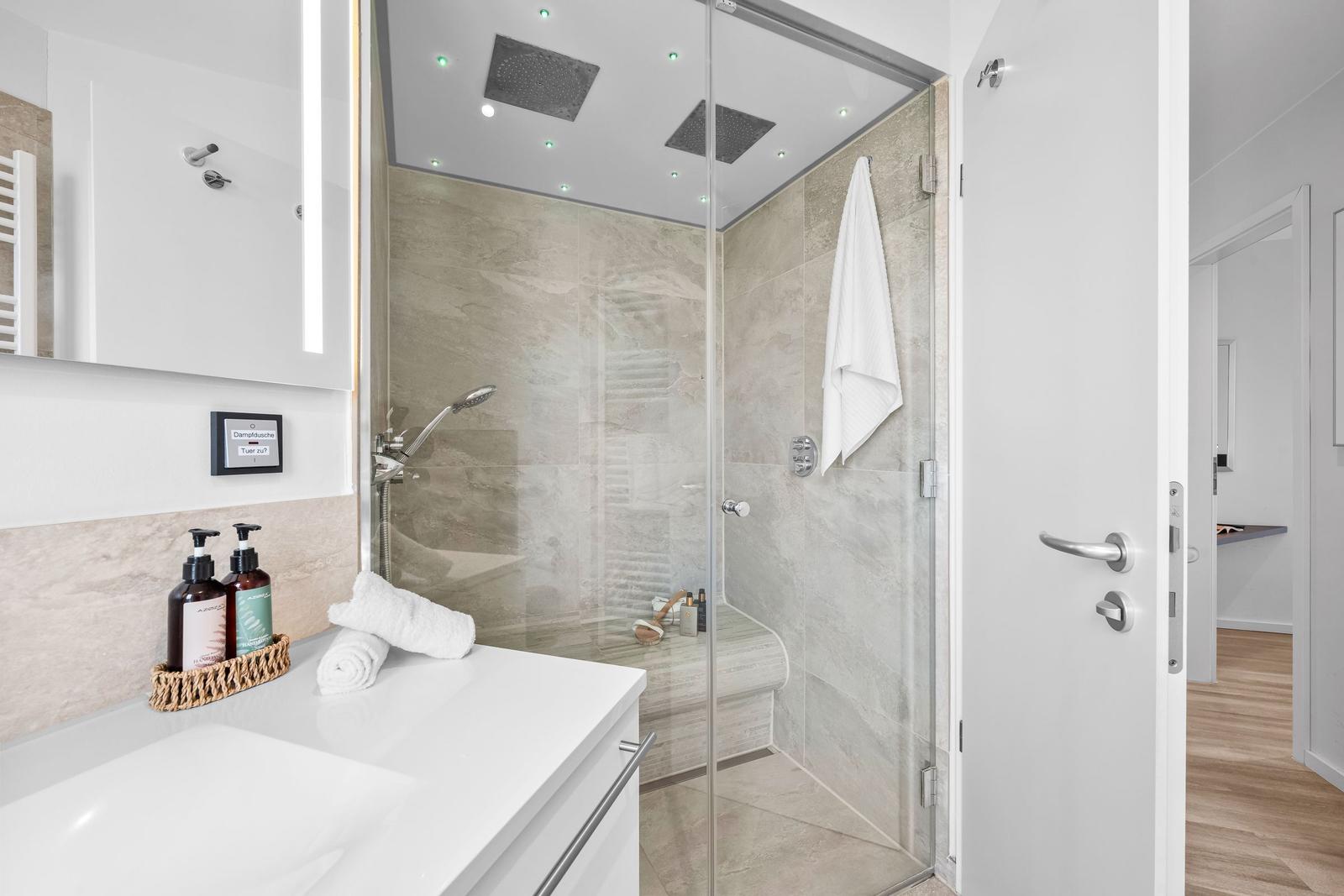 Moderne Dusche mit Glaswand, Duschbrause und Handtuch. Waschbecken mit Seife und Spiegel.