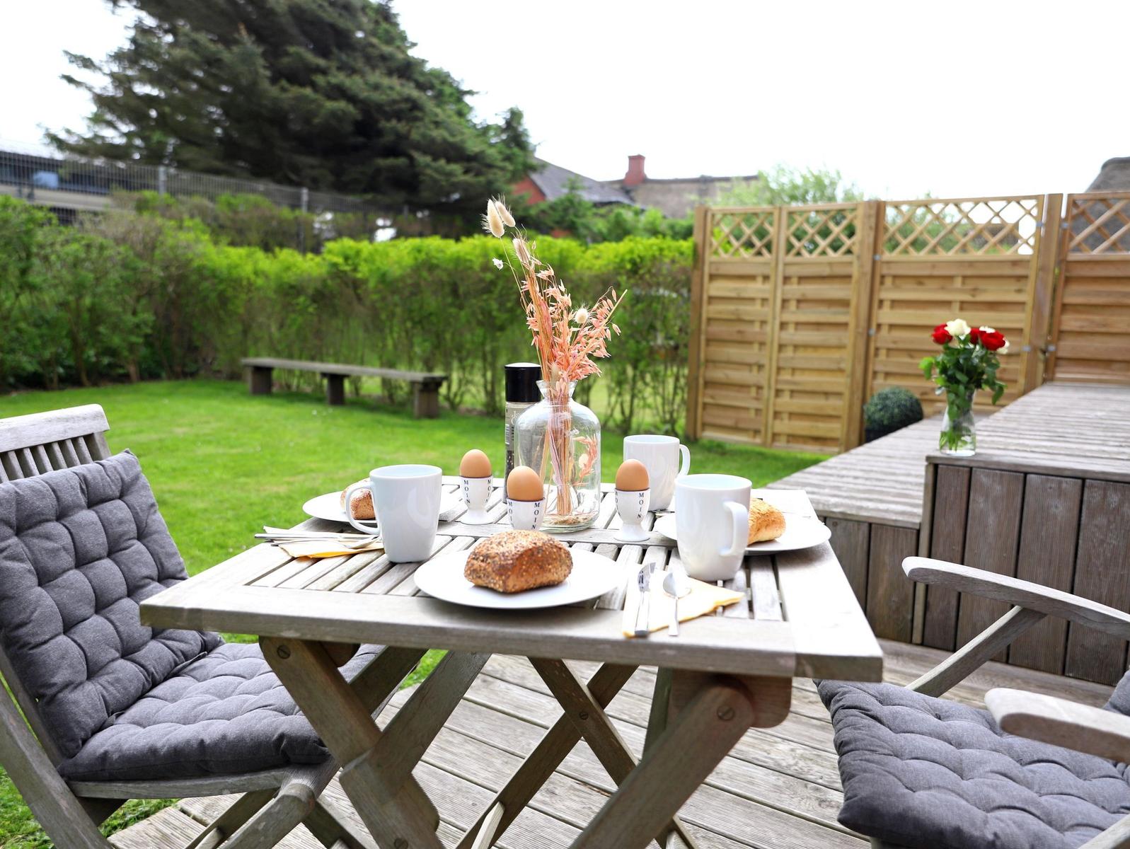 Holztisch mit Frühstück auf Terrasse, Hintergrund Grünfläche und Holzzaun.