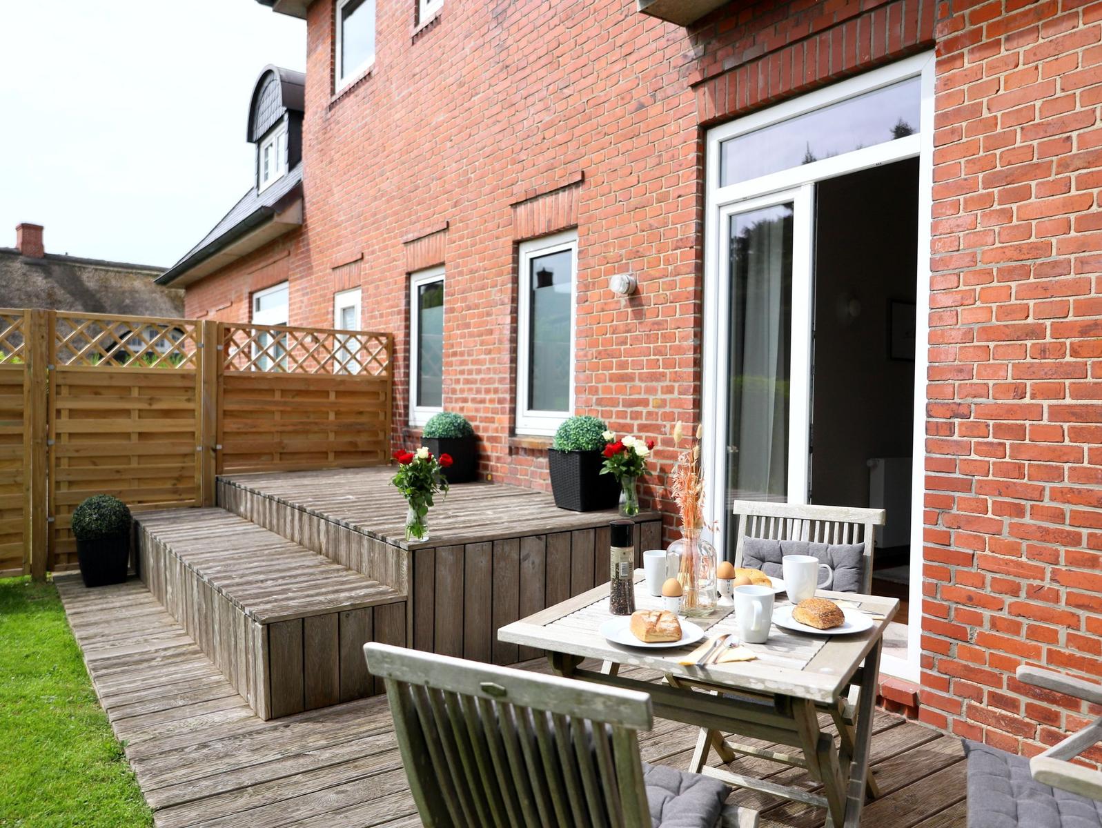 Holzterrasse mit Tisch, Stühlen und Blumen vor rotem Backsteinhaus.
