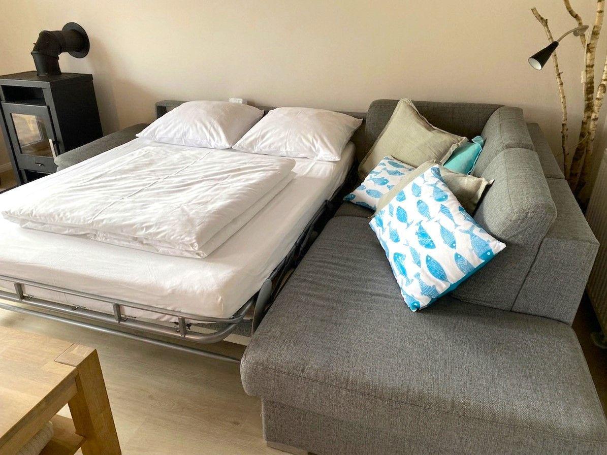 Schlafzimmer mit Bett und Sofa, daneben Holztisch und Kaminofen.