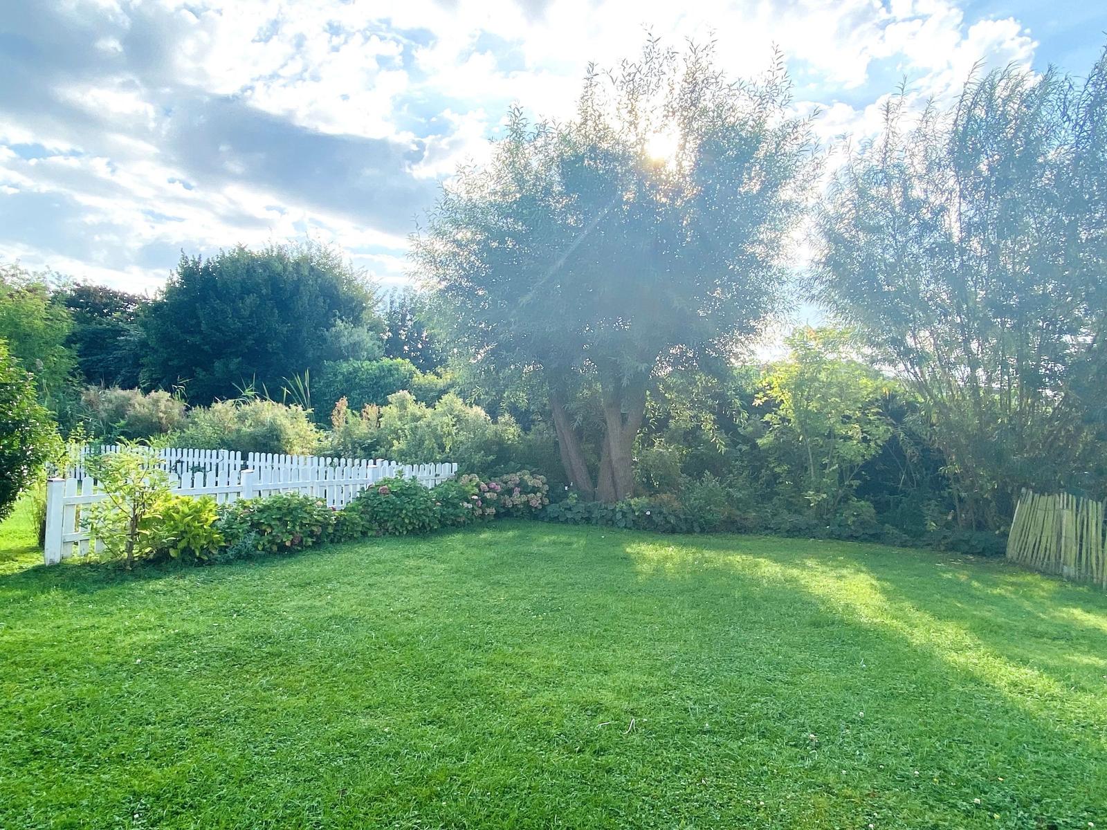 Garten mit grünem Rasen, weißen Zaun und Bäumen unter sonnigem Himmel.