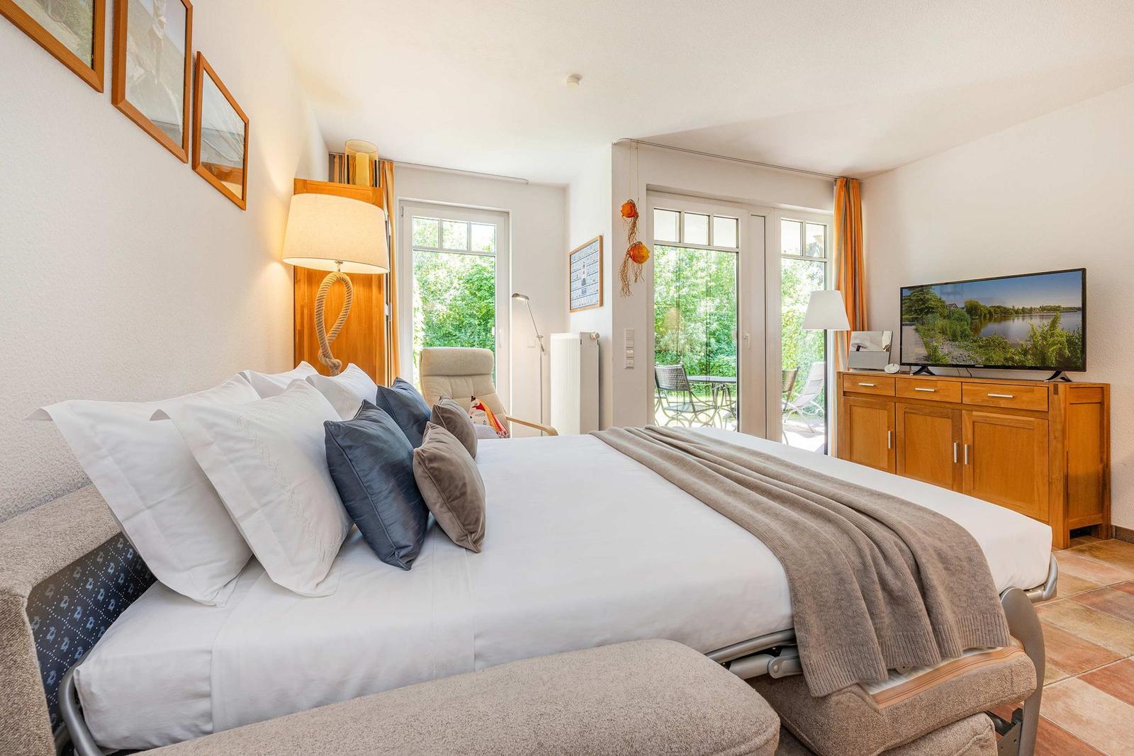 Schlafzimmer mit Bett, TV und Terrasse. Fenster und Tür führen nach draußen.