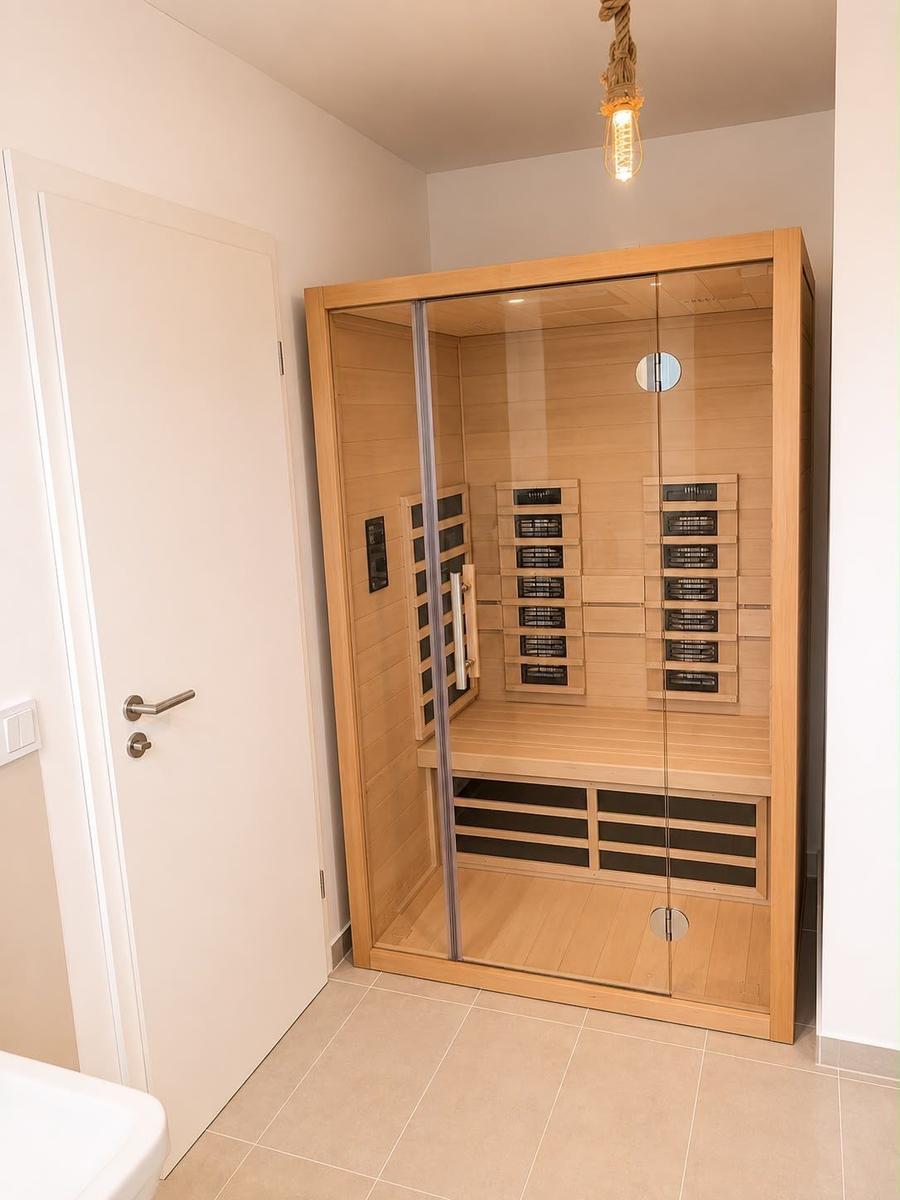 Innenraum einer Sauna mit Holzwand und Glasfront. Deckenlampe und Tür sind sichtbar.