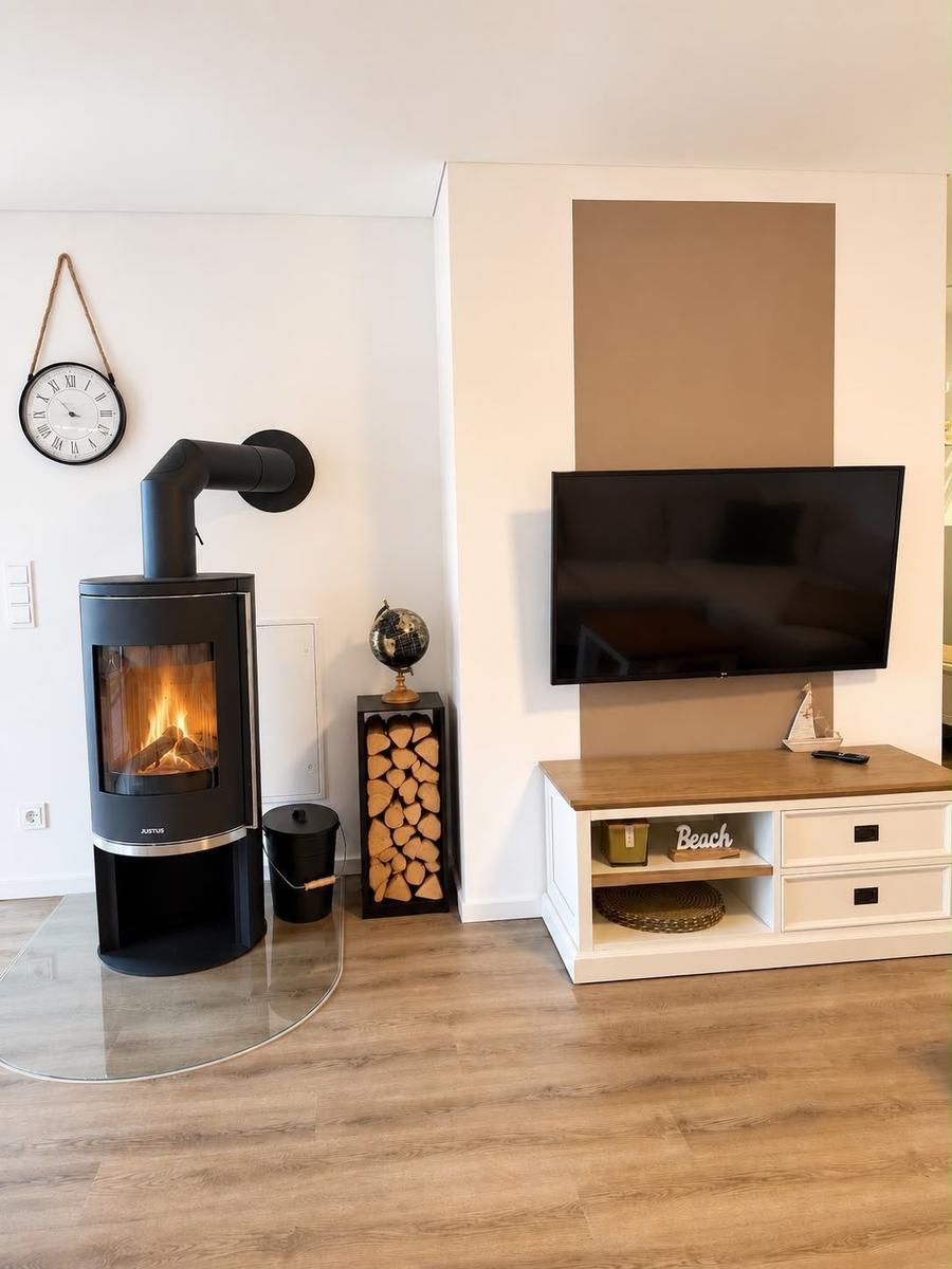 Wohnzimmer mit Kamin, Fernseher und Holzstapel. Wanduhr und Regal sichtbar.