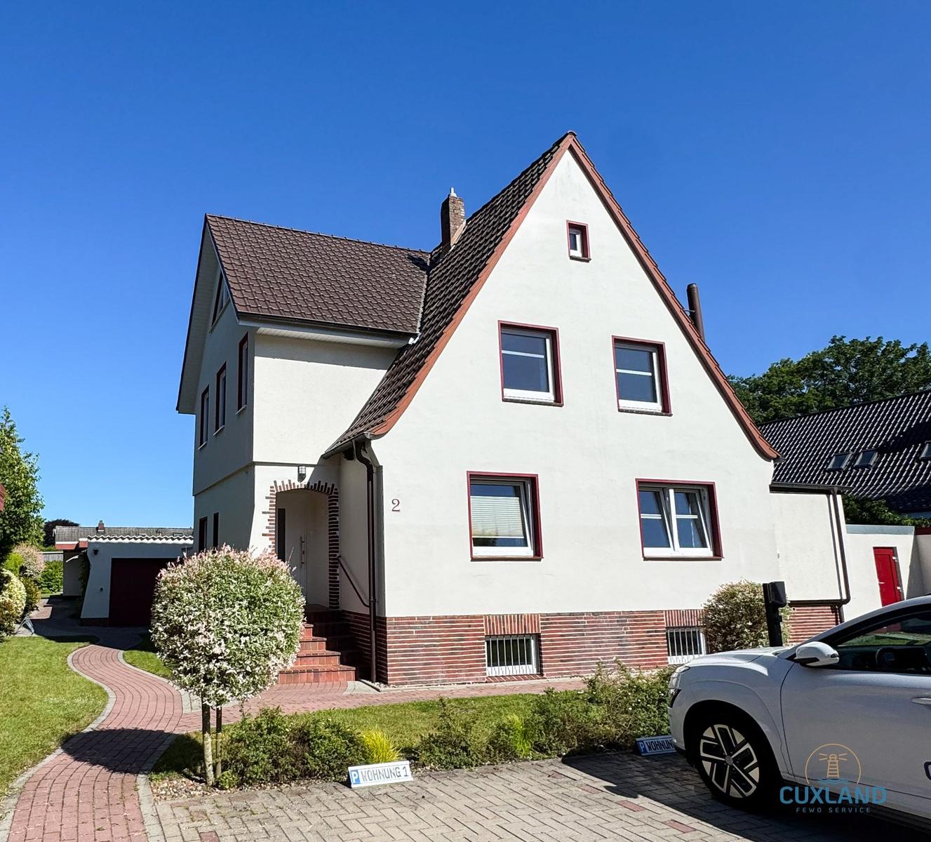Wohnhaus mit braunem Dach, weißen Wänden und roten Fensterrahmen. Vorhof mit gepflastertem Weg und gepflegtem Grün.