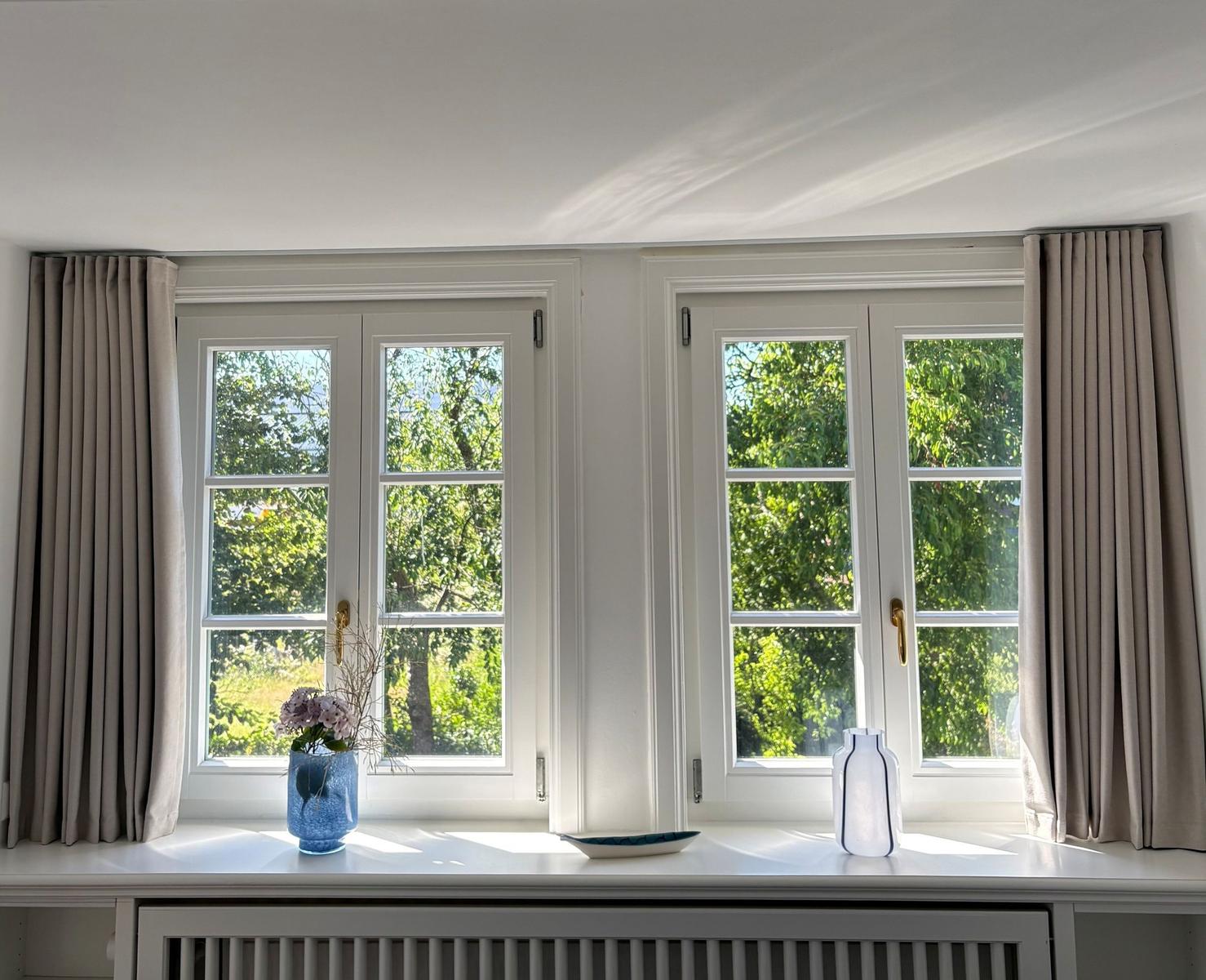 Zwei Fenster mit grauen Vorhängen und Blumen auf dem Fensterbrett.