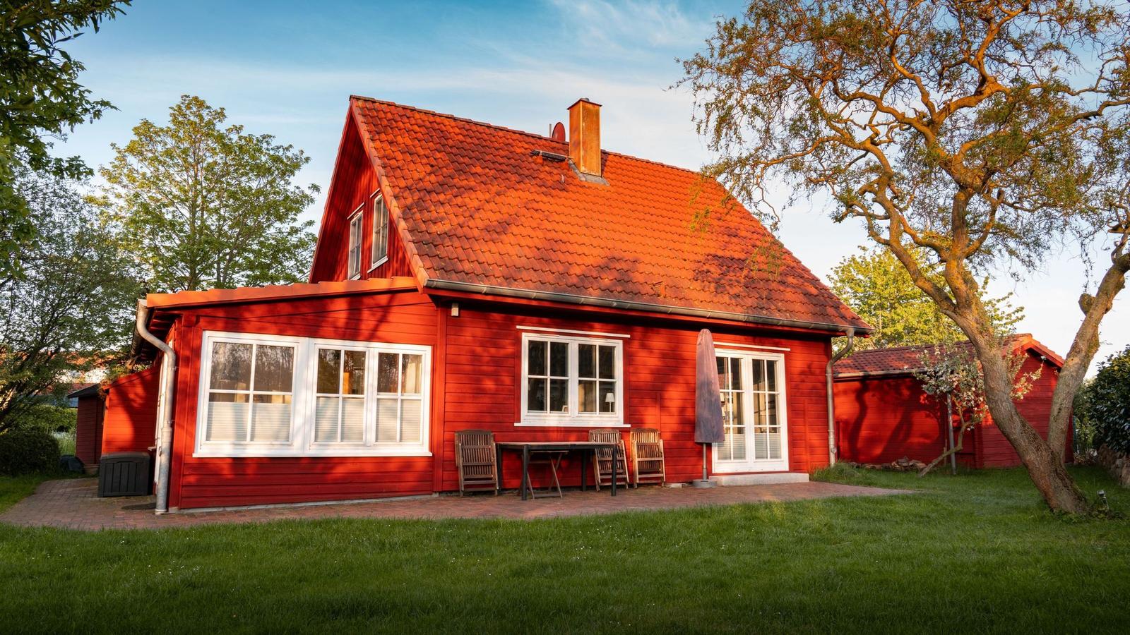 Rotes Holzhaus mit Terrasse, Tisch, Stühlen und großen Fenstern. Umgeben von Gras und Bäumen.