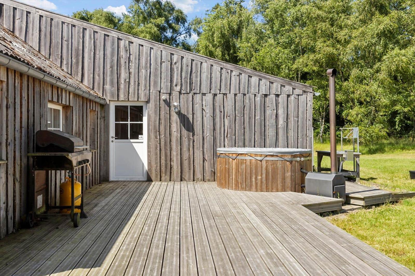 Holzterrasse mit Whirlpool, Grill und rustikalem Haus. Grün hinter dem Gebäude.