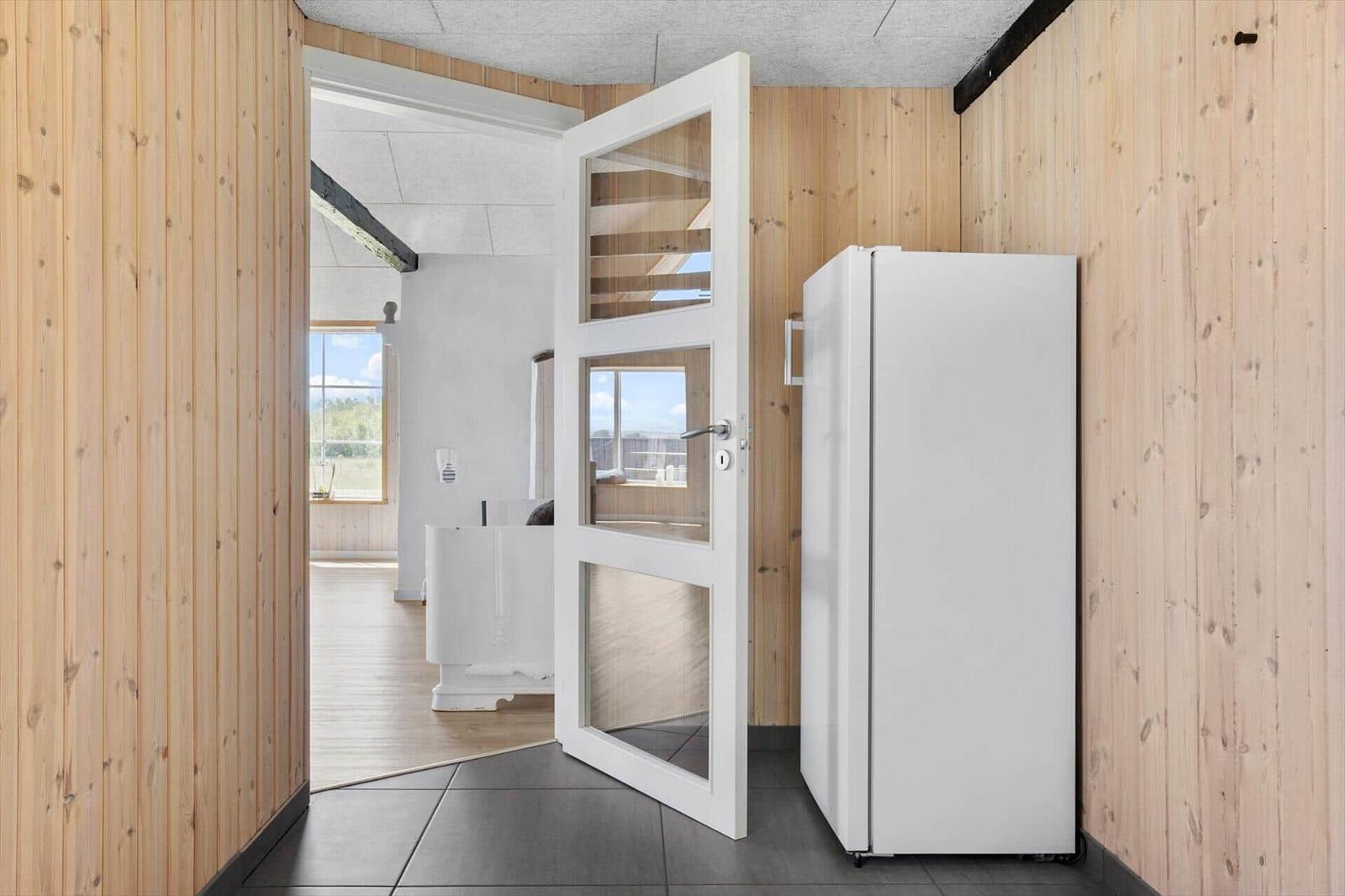 Küche mit weißem Kühlschrank und Holzwänden. Tür führt in Wohnbereich mit Fenster.