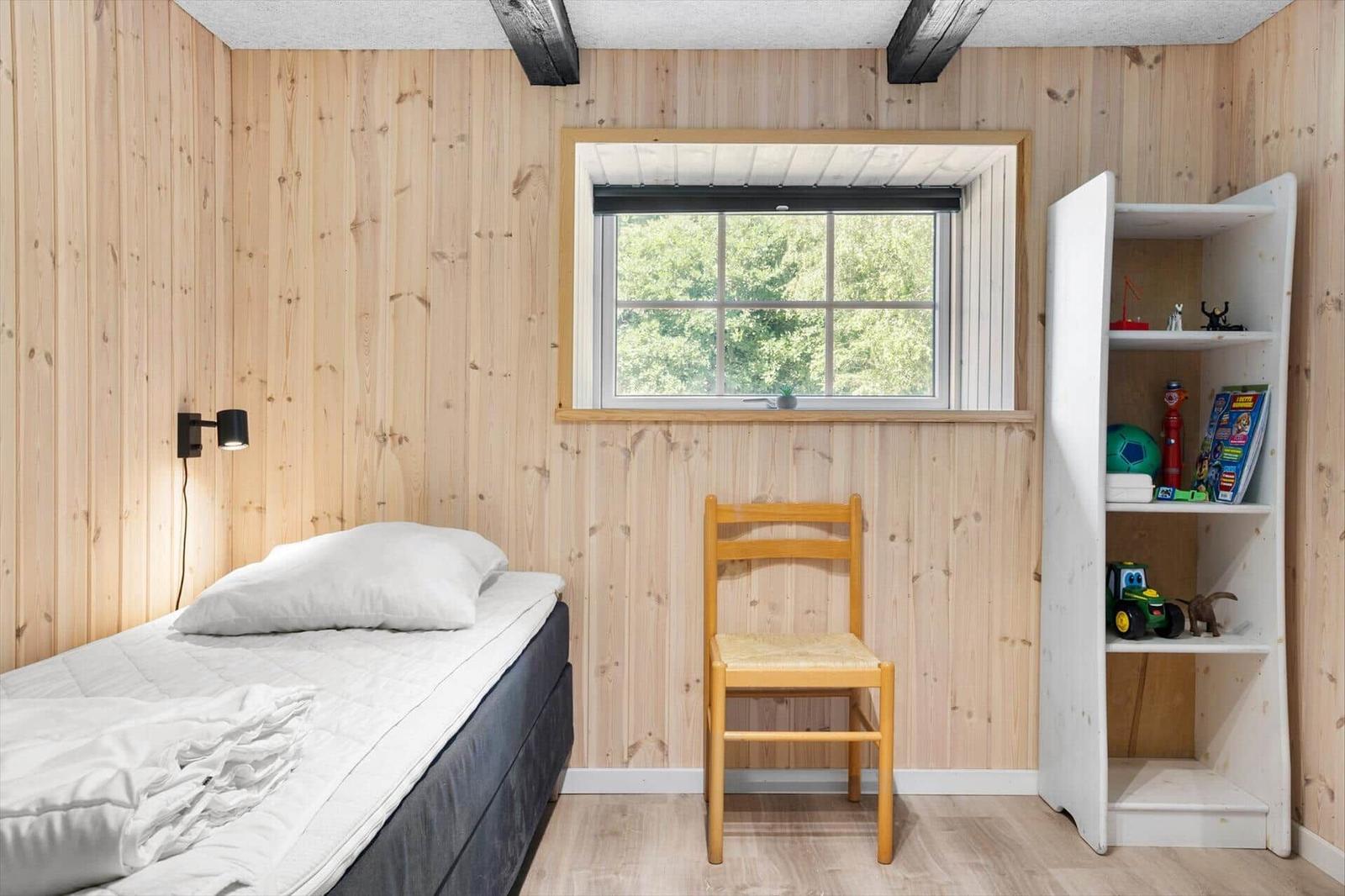 Ein Schlafzimmer mit Holzwand, Bett, Stuhl und Bücherregal. Fenster zeigt Wald.