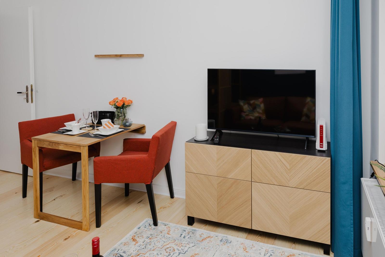 Ein Essbereich mit Holztisch, zwei orangefarbenen Stühlen und Fernseher auf einem Schrank.