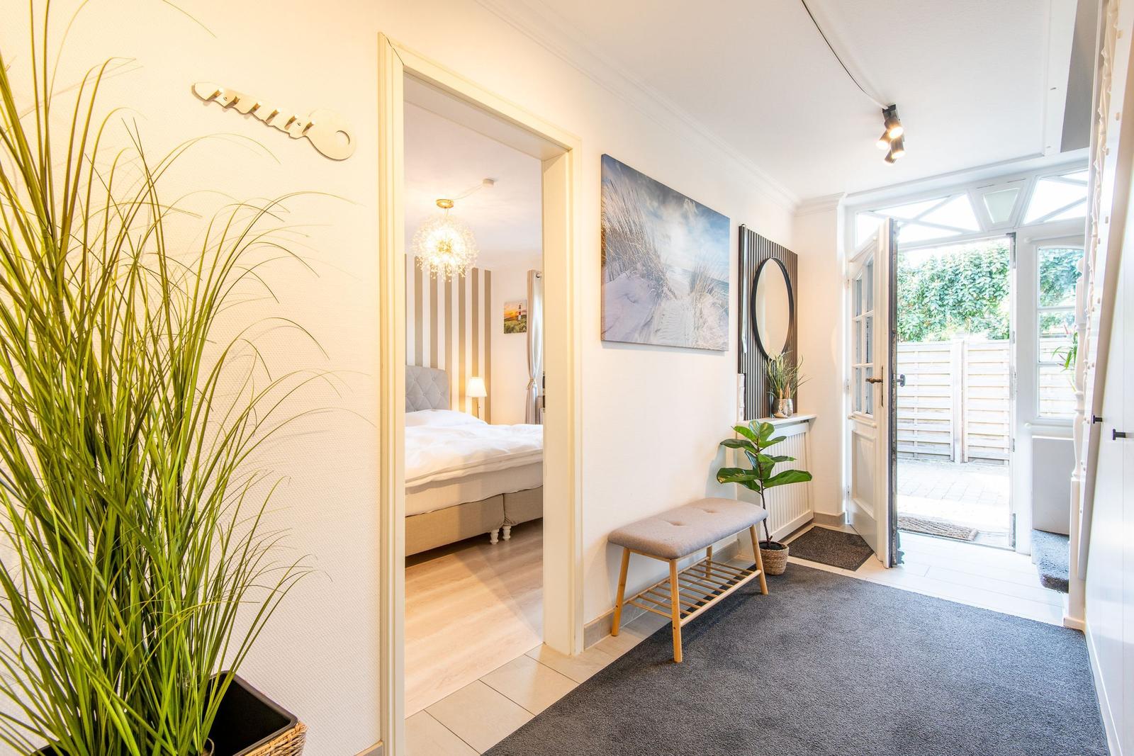 Eingangsbereich mit Sitzbank, Pflanzen und Blick in Schlafzimmer mit Bett und Deckenleuchte.