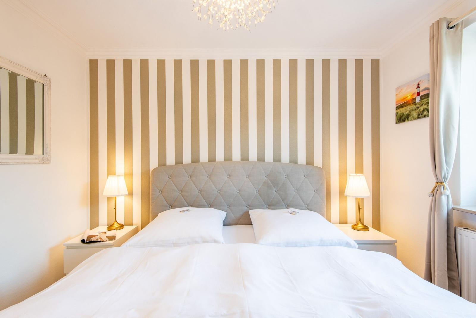 Schlafzimmer mit gestreifter Wand, Bett, Nachttischen und Lampen.
