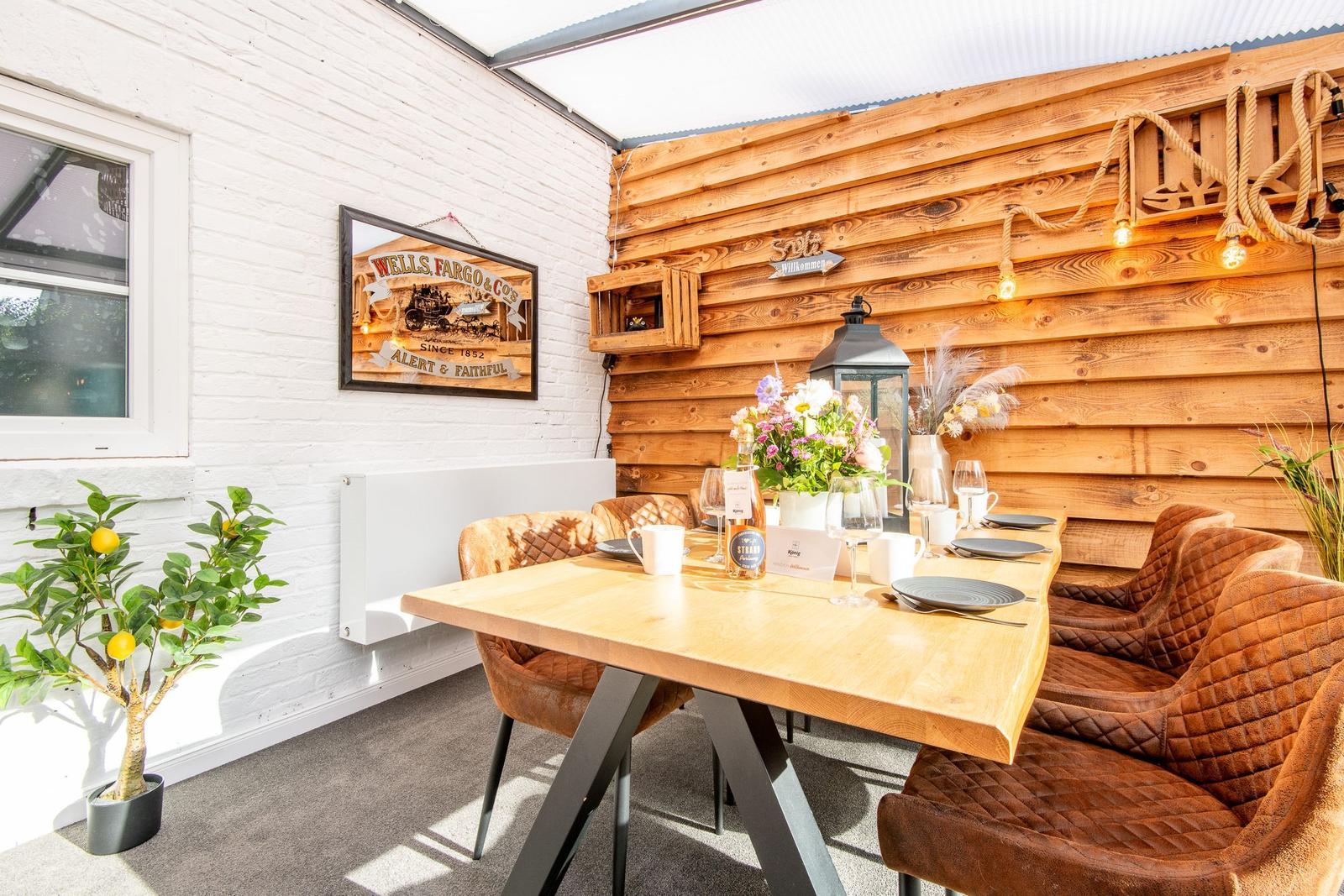Terrassenbereich mit Esstisch, Stühlen und Blumenarrangement. Holzwand und weiße Wand mit Bild.