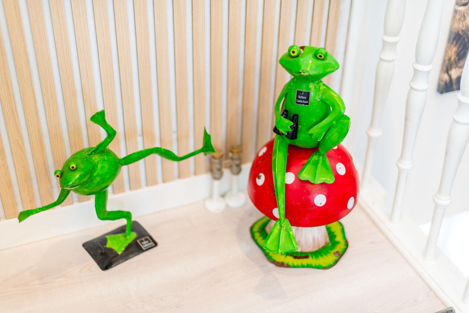 Zwei grüne Froschfiguren, eine springt, die andere sitzt auf einem roten Pilz.