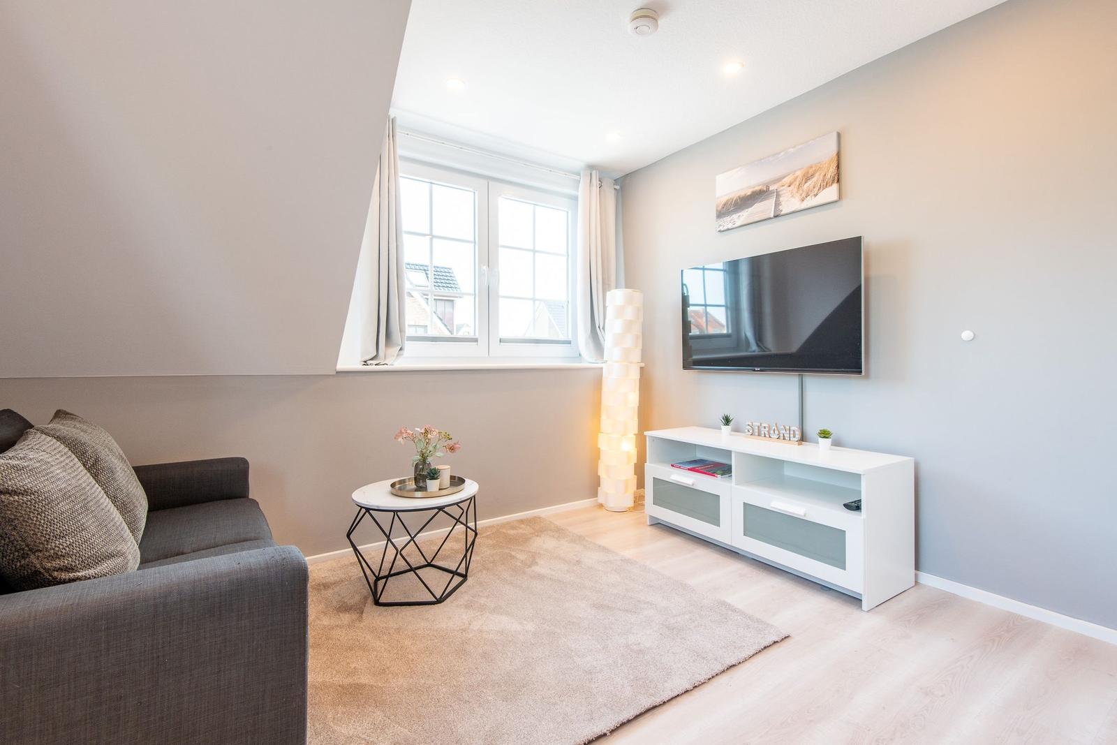 Wohnzimmer mit Sofa, TV und Fenster. Flur und Teppich sind sichtbar.