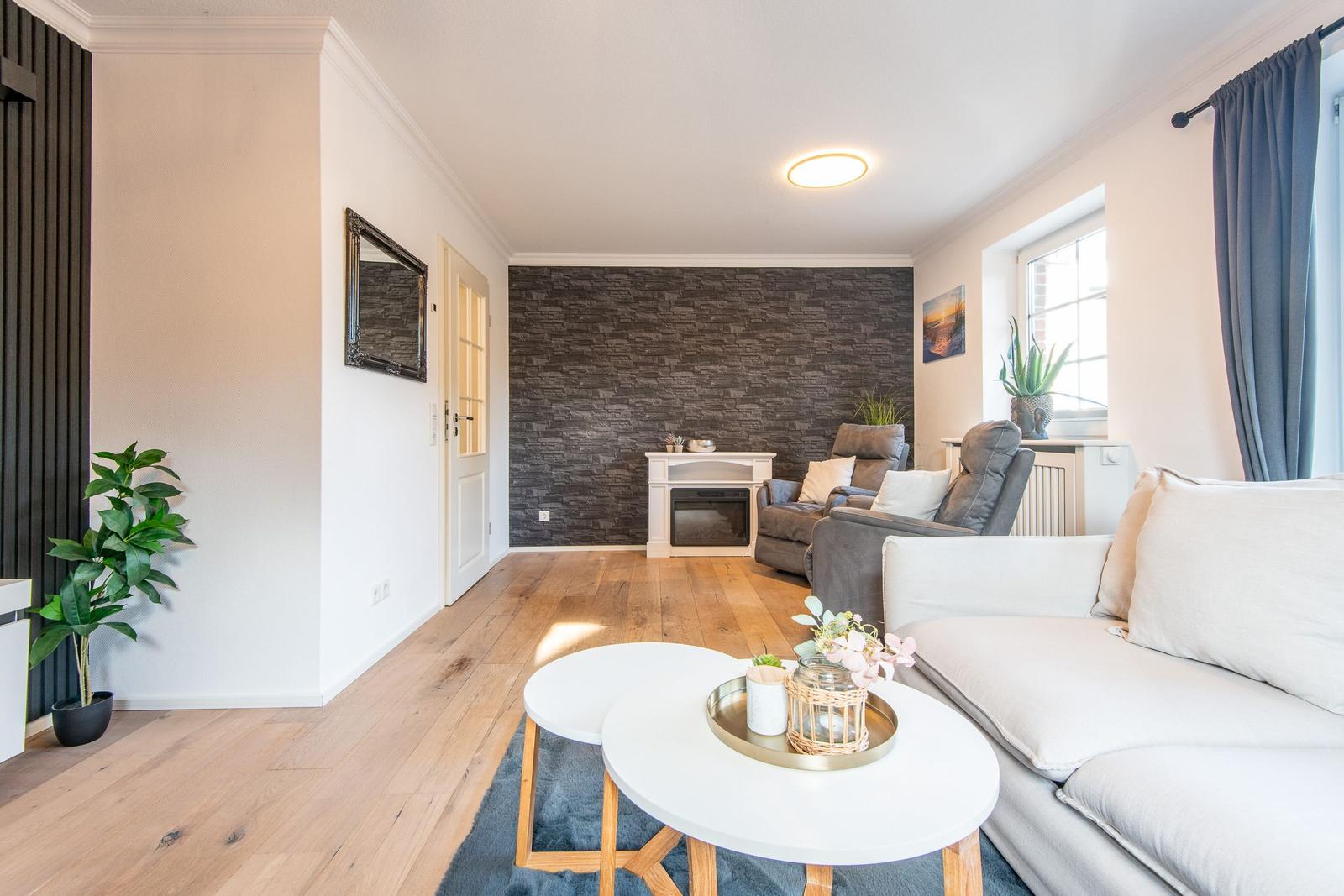 Wohnzimmer mit Sofa, Sitzgelegenheit und Holzboden. Wand mit Steinoptik und Fenster mit Vorhang.
