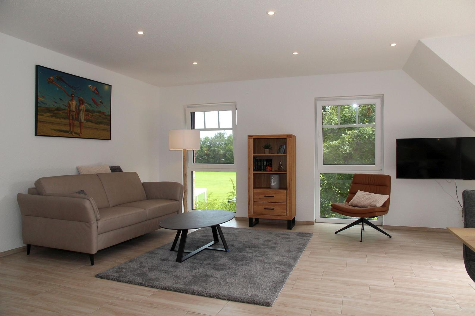 Wohnzimmer mit Sofa, Sessel, Tisch und Fenstern. Wandbild und TV sichtbar.