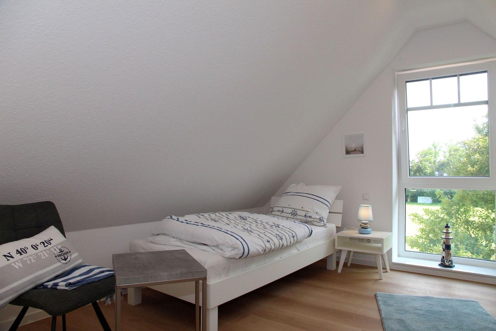 Schlafzimmer mit Dachschräge, Bett, Stuhl und großem Fenster mit Blick auf Grünfläche.