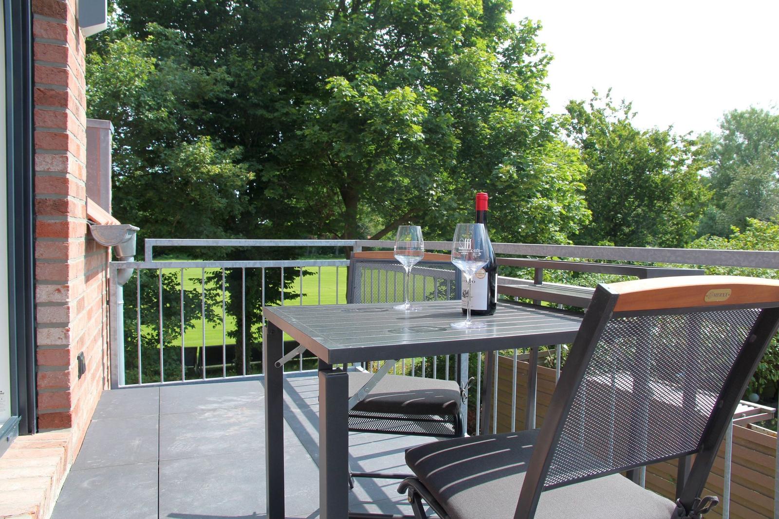 Terrasse mit Tisch, zwei Stühlen, Weinflasche und Gläsern. Blick auf grüne Wiesen und Bäume.