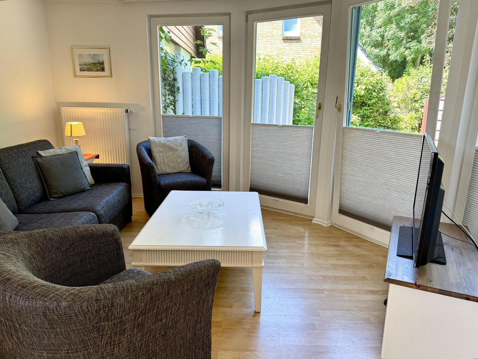 Wohnzimmer mit Sofa, Sessel, Tisch und Fernseher. Durchgänge zu einem Garten.