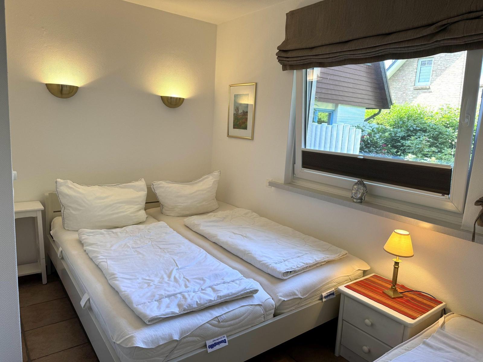Zimmer mit zwei Betten, Fenster und Nachttisch mit Lampe.